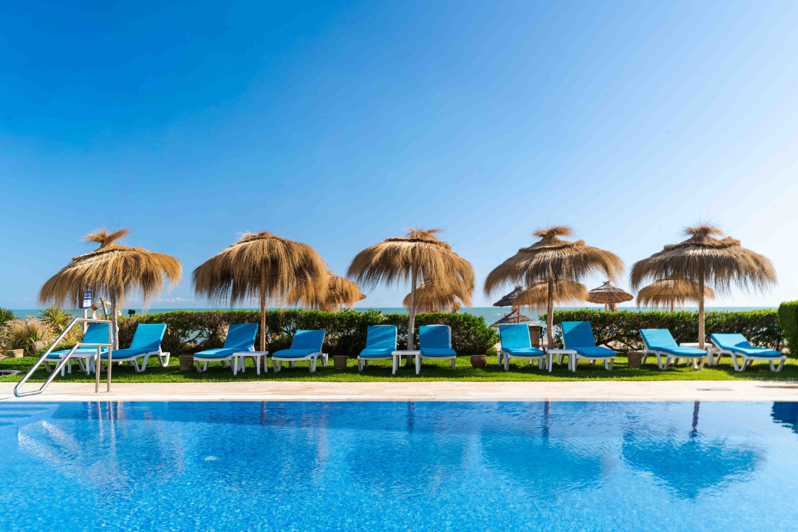 Piscine avec transats bleus et parasols en paille face à la mer